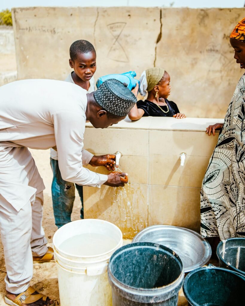 Villagers gather at a communal faucet to collect water on a sunny day.
